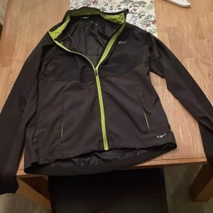 Svart jacka från SOC - Säljer en svart jacka från SOC med limegröna detaljer och dragkedja. Jackan har en sportig design med långärmade ärmar och är perfekt för utomhusaktiviteter. Den har en bekväm passform och är tillverkad i ett syntetmaterial som ger bra skydd mot vind. Aldrig använt 