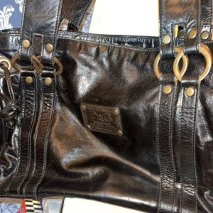 Svart skinnväska från Dolce & Gabbana - Snygg svart skinnväska från Dolce & Gabbana med dekorativa ringdetaljer och nitar. Väskan har dubbla handtag och en praktisk innerficka med dragkedja. Perfekt för att bära med sig det nödvändigaste med stil.