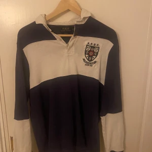 Randig pikétröja från Ralph Lauren - Säljer en stilren randig pikétröja från Ralph Lauren i blått och vitt. Tröjan har långa ärmar och en klassisk krage med knappar. Perfekt för en avslappnad men ändå snygg look.