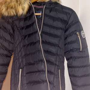Svart pufferjacka, vinson polo club - Snygg svart pufferjacka med dragkedja och huva med pälsdetalj. Jackan har långa ärmar och praktiska fickor med dragkedjor. Perfekt för kyliga dagar.