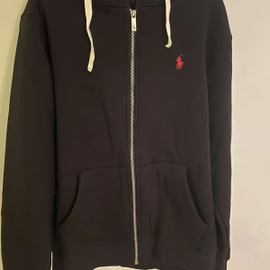 Svart hoodie från Ralph Lauren - Snygg svart hoodie från Ralph Lauren med dragkedja och vit snörning. Den har en klassisk design med det ikoniska röda logotypen på bröstet. Perfekt för en avslappnad stil.