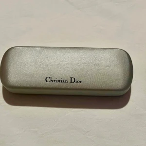 Glasögonfodral från Christian Dior - Elegant glasögonfodral från Christian Dior i en stilren silverfärg. Fodralet har en klassisk design med logotypen tryckt på utsidan och insidan. Perfekt för att skydda dina glasögon med stil. Några små skavmärken finns 
