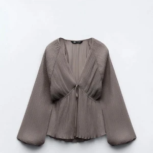 Grå blus från Zara - Elegant grå blus från Zara med v-ringning och knytning framtill. Blusen har vida, långa ärmar och ett plisserat tyg som ger en stilren look. Perfekt för både vardag och fest.