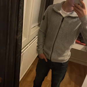 Grå stickad tröja från Sail Antarctica - Säljer en snygg grå stickad tröja från Sail Antarctica. Den har en dragkedja framtill och en cool patch på ärmen. Perfekt för kyliga dagar och passar bra till både jeans och chinos. Tröjan är i mycket bra skick och redo för nya äventyr!