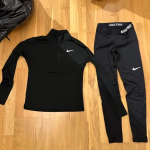 Svarta Nike Pro leggings och matchande tröja - Säljer ett par svarta Nike Pro leggings och en matchande tröja i bra skick. De är perfekta för träning med sin tighta passform och elastiska midja. Materialet är mjukt och stretchigt, vilket ger bra rörelsefrihet. Nike-loggan syns tydligt på benet, midjan och bröstet.