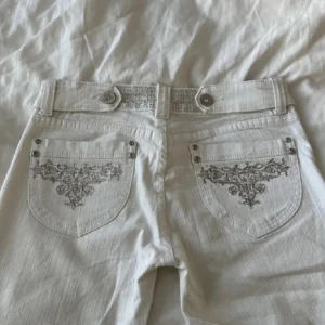 Vita lowwaist jeans med broderi - Säljer ett par liknande snygga vita jeans som på bilden fast det är knappfickor som true religion jeans på bakfickorna och framsidorna💗(hör av dig för bilder). De har en bootcut-stil och är dekorerade med knappar och detaljer vid midjan. Aldrig använda!