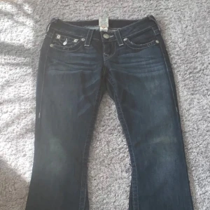 Mörkblå jeans med broderade fickor - Säljer ett par snygga mörkblå jeans från True Religion. De har coola broderade fickor och en klassisk femficksdesign. Jeansen är i bra skick och har en bootcut-stil som ger en avslappnad look. Perfekta för både vardag och fest!   Orginalpris 1300
