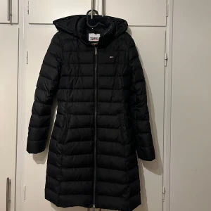 Svart dunjacka från Tommy Jeans - Säljer en svart dunjacka från Tommy Jeans i storlek S. Jackan har en lång figursydd-modell med dragkedja och huva, perfekt för kalla vinterdagar. Den är i mycket bra skick och har en stilren design med märkets logga på bröstet och ärmen.