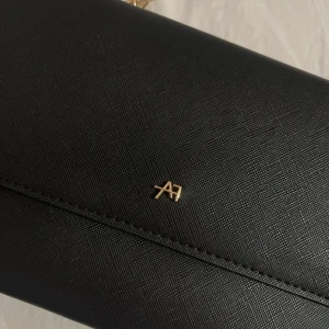 Svart kuvertväska med guldkedja - Snygg svart kuvertväska i syntetmaterial med en elegant guldkedja som axelrem. Perfekt för en utekväll eller fest. Väskan har en stilren design med en liten logotypdetalj i guld på framsidan. Enkelt och stilrent val för att komplettera din outfit.