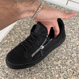 giuseppes zanotti - Snygga svarta sneakers i syntetmaterial med coola dragkedjor på sidan. De har en glittrig yta som ger en extra touch av stil. Perfekta för både vardag och fest! Skorna har snörning och en bekväm passform.