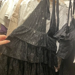Svart glittrig klänning - Säljer en superfin svart klänning med glittriga detaljer. Den har en design som gör att man knyter den runt nacken och volanger som ger en festlig look. Perfekt för nyår eller andra festliga tillfällen. Klänningen är i bra skick och har en bekväm passform. Vart den kommer ifrån är okänt.