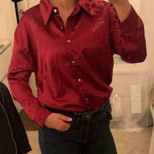 Röd satinblus - Snygg röd satinblus med långa ärmar och knappar framtill. Perfekt för festliga tillfällen eller en utekväll. Blusen har en klassisk krage och en glansig finish som ger en lyxig känsla. Passar bra till både jeans och kjol.