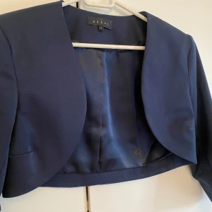 Mörkblå fest kavaj bolero från Debut London - Säljer en elegant mörkblå kavaj från Debut London. Den är kort och stilren, perfekt för formella tillfällen. Kavajen har en öppen design utan knappar och långa ärmar. Passar bra till både klänning och byxor. Bolero till fest. 