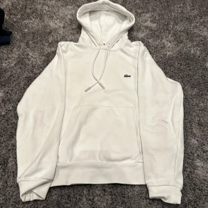 Vit hoodie från Lacoste - Säljer en stilren vit hoodie från Lacoste i klassisk passform. Den har en känguruficka och justerbar huva med dragsko. Perfekt för en avslappnad stil och passar till alla säsonger. Riktigt skön. 🐊