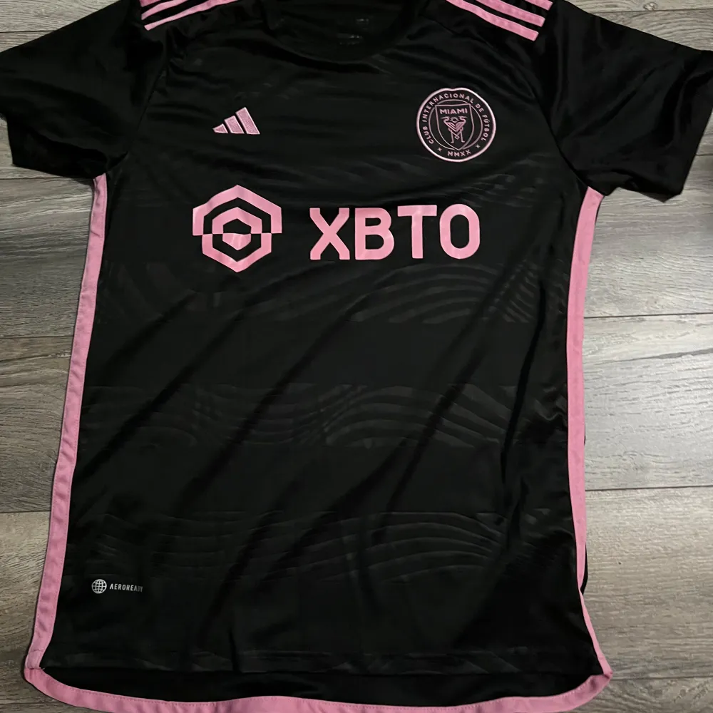 Säljer en snygg svart fotbollströja med rosa detaljer från Adidas. Tröjan har korta ärmar och Messi 10 tryckt på ryggen. Perfekt för fotbollsfans som vill ha en stilren look på planen eller på stan. Materialet är lätt och andas bra. Passar både till träning och vardag.. T-shirts.