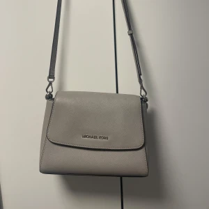 Grå axelväska från Michael Kors - Säljer en stilren grå axelväska från Michael Kors i mycket bra skick. Väskan har en justerbar axelrem och ett snyggt, strukturerat material. Knappt använd då jag mest använder guldsmycken vilket gör att den inte passar till. Köpt i USA i Michael Kors egna butik 