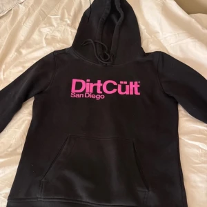 Svart hoodie från DirtCult - Säljer en svart hoodie från DirtCult med rosa tryck 'San Diego' på bröstet. Den har en klassisk känguruficka och justerbar huva. Perfekt för en avslappnad stil eller kyliga dagar. 🖤💖tröjan är i L men passar även mig som har s/xs
