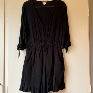 Svart byxdress - Säljer en elegant svart byxdress med trekvartsärmar och knytband vid ärmarna. Byxdressen har en v-ringad design och elastisk midja för en bekväm passform. Perfekt för både vardag och festliga tillfällen!