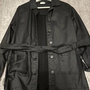 Svart kappa från VILA - Snygg svart kappa från VILA i mycket bra skick. Den har en klassisk krage och knappar framtill. Jackan är långärmad och har ett bälte i midjan för en elegant look. Perfekt för höst och vår!