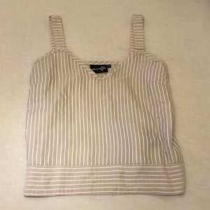 Beige och vit randig linnetopp från Pelle P - Säljer en superfin beige och vit randig linnetopp från Pelle P. Perfekt för sommardagar med sin lätta och luftiga design. Toppen har breda axelband och en rak passform som gör den både bekväm och stilren. Passar perfekt till både jeans och kjol!