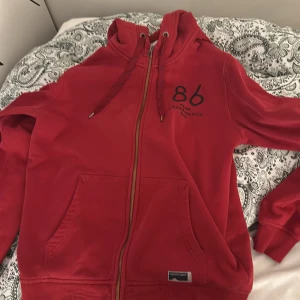 Röd hoodie från Peak Performance - Säljer en snygg röd hoodie från Peak Performance med dragkedja och justerbar huva. Den har en cool '86' design på bröstet och är perfekt för både vardag och chill. Superbekväm och i bra skick!