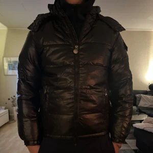 Svart pufferjacka - Säljer en svart pufferjacka i mycket bra skick, då den är endast upp packad. Den är perfekt för kalla dagar med sin vadderade design och dragkedja framtill. Jackan har långa ärmar och en hög krage för extra värme. Passar bra till både vardag och vinteraktiviteter.