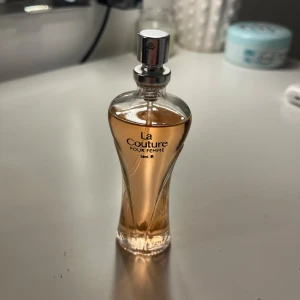 La Couture Pour Femme Parfym - Säljer en elegant parfymflaska från La Couture, Pour Femme. Flaskan har en smal och kurvig design med en lyxig känsla. Den är genomskinlig med en ljusrosa vätska inuti och en silverfärgad spraytopp. Perfekt för en sofistikerad doftupplevelse.