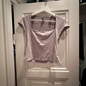 Grå kortärmad topp från H&M - Säljer en stilren grå topp från H&M. Den har korta ärmar och en fyrkantig halsringning som ger en modern look. Perfekt för både vardag och festliga tillfällen. Toppen är i bra skick och passar till det mesta i garderoben. Skrynklig pga legat i påse på vinden