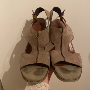 Beige mockasandaler från Tamaris - Snygga beige sandaler i mocka från Tamaris med en stabil klack och öppen tå. De har en elegant guldfärgad spänne och är perfekta för sommarens alla fester. Skorna är i mycket bra skick och har en bekväm passform.