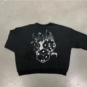 Säljer en svart sweatshirt från Stussy med ett coolt tryck av tärningar och eldflammor på ryggen. Perfekt för en avslappnad stil. Tröjan har långa ärmar och är i mycket bra skick.