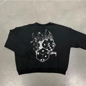 Svart sweatshirt från Stussy med tärningsmotiv - Säljer en svart sweatshirt från Stussy med ett coolt tryck av tärningar och eldflammor på ryggen. Perfekt för en avslappnad stil. Tröjan har långa ärmar och är i mycket bra skick.