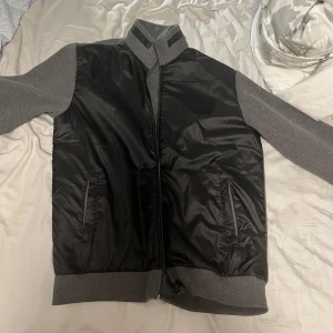 Svart och grå, Hugo boss cardigan - Säljer en snygg svart och grå bomberjacka med dragkedja. Jackan har långa ärmar och är perfekt för höst och vår. Den är i bra skick och har en stilren design med ribbade muddar och krage. Perfekt för en casual look!