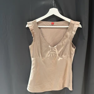 Beige sidenlinne med volangdetaljer - Elegant beige linne i siden med volangdetaljer vid axlarna och en v-ringad design. Perfekt för en stilren och sofistikerad look.