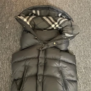 Svart dunväst med rutigt foder - Snygg svart dunväst med dragkedja och knappar. Insidan har ett stilrent rutigt mönster i grått och svart. Perfekt för kyligare dagar med sin quiltade design.