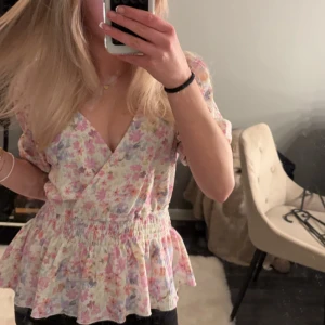 Blommig omlottblus - Säljer en söt blommig blus med volangdetaljer. Blusen har en v-ringad design och korta ärmar. Perfekt för vår och sommar!