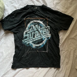 Svart t-shirt med The Strokes-tryck från Pull&Bear - Cool svart t-shirt från Pull&Bear med ett stort The Strokes-tryck i retrostil på framsidan. Perfekt för fans av bandet eller för en rockig look. Tillverkad i mjukt material för bekväm passform. Klicka gärna på köp nu