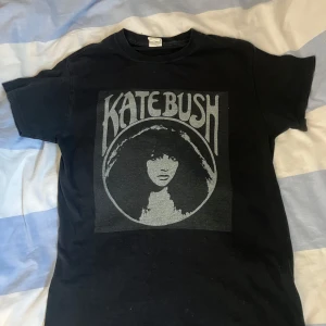Svart t-shirt med Kate Bush-tryck - Säljer en svart t-shirt med ett stort tryck av Kate Bush i grått på framsidan. T-shirten har en klassisk passform och korta ärmar. Använd men utan defekter. Storlek S och i 100% bomull. 