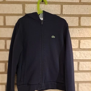 Mörkblå hoodie från Lacoste Sport - Säljer en snygg mörkblå hoodie från Lacoste Sport med dragkedja och den klassiska krokodilloggan på bröstet. Perfekt för en sportig look eller en avslappnad dag. Den har en bekväm huva och långa ärmar.