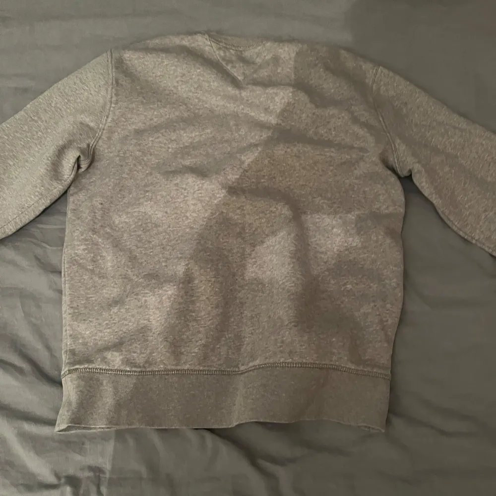 Säljer en klassisk grå sweatshirt från Tommy Hilfiger med logotyp och text 'EST. 1985' på framsidan. Tröjan har rund hals och långa ärmar, perfekt för en avslappnad stil. Den är mjuk och bekväm, idealisk för vardagsbruk.. Neuletakit & Villapaidat.