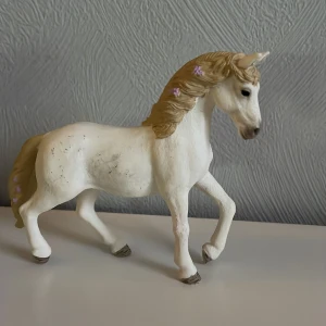 Schleich häst - Charmig vit hästfigur med detaljerade blommor i manen. Hästen har en elegant pose och är perfekt för samlare eller som dekoration. Manen och svansen är ljusbruna med små lila blommor som ger en fin touch.
