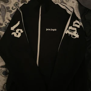 Svart kofta från Palm Angels - Säljer en svart kofta från Palm Angels med vita detaljer och dragkedja. Jackan har långa ärmar med tryck i vitt på ärmarna och bröstet. Perfekt för en sportig look!