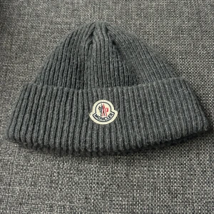 Grå ribbad mössa från Moncler - Snygg grå ribbad mössa från Moncler med broderad logotyp framtill. Perfekt för kyliga dagar och ger en stilren look. Tillverkad i ett mjukt och bekvämt material godtas hälst av byten men den sälj även!