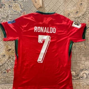 Portugal Fotbollströja Ronaldo  - Säljer en röd fotbollströja från Nike med nummer 7 och namn på baksidan. Tröjan har korta ärmar och gröna detaljer vid ärmslut och krage. Perfekt för fotbollsfans!