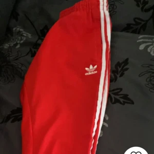 Röda träningsbyxor från Adidas - Snygga röda träningsbyxor från Adidas med de klassiska tre vita ränderna längs sidorna. Byxorna har en elastisk midja och dragkedja vid fickorna för extra funktionalitet. Perfekta för en sportig look eller träning. Använd några gånger men är ändå i bra skick 