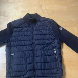  Moncler jacka - Snygg svart pufferjacka från Moncler med dragkedja och ribbad krage. Jackan har långa ärmar och två praktiska fickor med dragkedja framtill. Perfekt för kyligare dagar.