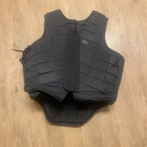 Svart skyddsväst från Rider's Company - Säljer en svart skyddsväst från Rider's Company. Västen har en quiltad design och justerbara sidoband för optimal passform. Perfekt för ridning och andra aktiviteter där extra skydd behövs. Använd 2 gånger så i ny skick.