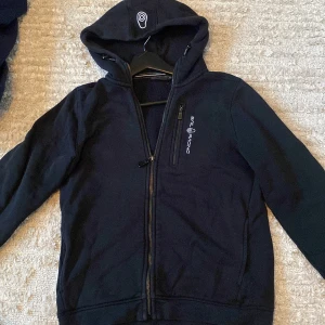 Svart zip hoodie från sail racing(160 cm) - Säljer en svart jacka från Sail Racing med dragkedja och huva. Jackan har en broderad logotyp på bröstet och huvan. Perfekt för kyligare dagar.