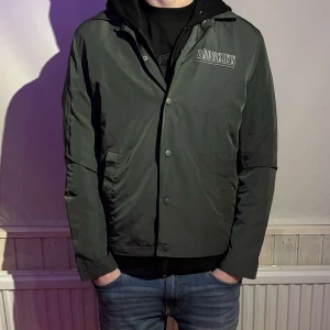 Grön jacka med Brooklyn-tryck - Snygg grön jacka med texten 'Brooklyn' tryckt på bröstet och 'Rockaway Ave. Brooklyn' på ryggen. Jackan har knappar framtill och är långärmad. Perfekt för en cool och avslappnad stil.