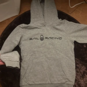 Grå hoodie från Sail Racing - Säljer en grå hoodie från Sail Racing med tryck på bröstet. Tröjan har en klassisk design med huva och ribbade muddar vid ärmslut och nederkant. Perfekt för en avslappnad stil.