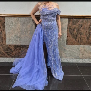 Ljusblå axelbandslös klänning - Elegant ljusblå axelbandslös klänning med glittriga detaljer och en dramatisk slits. Klänningen har en omlott-design och en lång, svepande kjol som ger en lyxig känsla. Perfekt för speciella tillfällen.